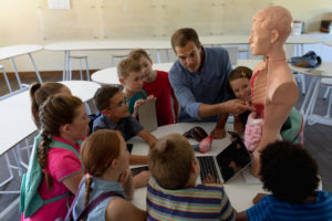 Front View Of A Caucasian Male Teacher Using A Human Anatomy Model To Teach A Diverse Group Of Elementary School Children During A Biology Lesson, The Children Sitting In A Circle And Listening While He Talks, Pointing To Organs On The Model