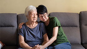 Supporting Persons Living With Dementia Graphic of 2 ladies sitting on a sofa. The younger lady is comforting the older lady.