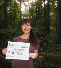 TCOM alumna Dr.Megan Jenkins-Turner stands in wooded trail holding a piece of paper that says, "I matched at UI Health in General Surgery Prelim"