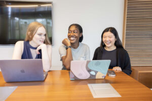 Photograph of three students smiling while sitting at a table working on laptops