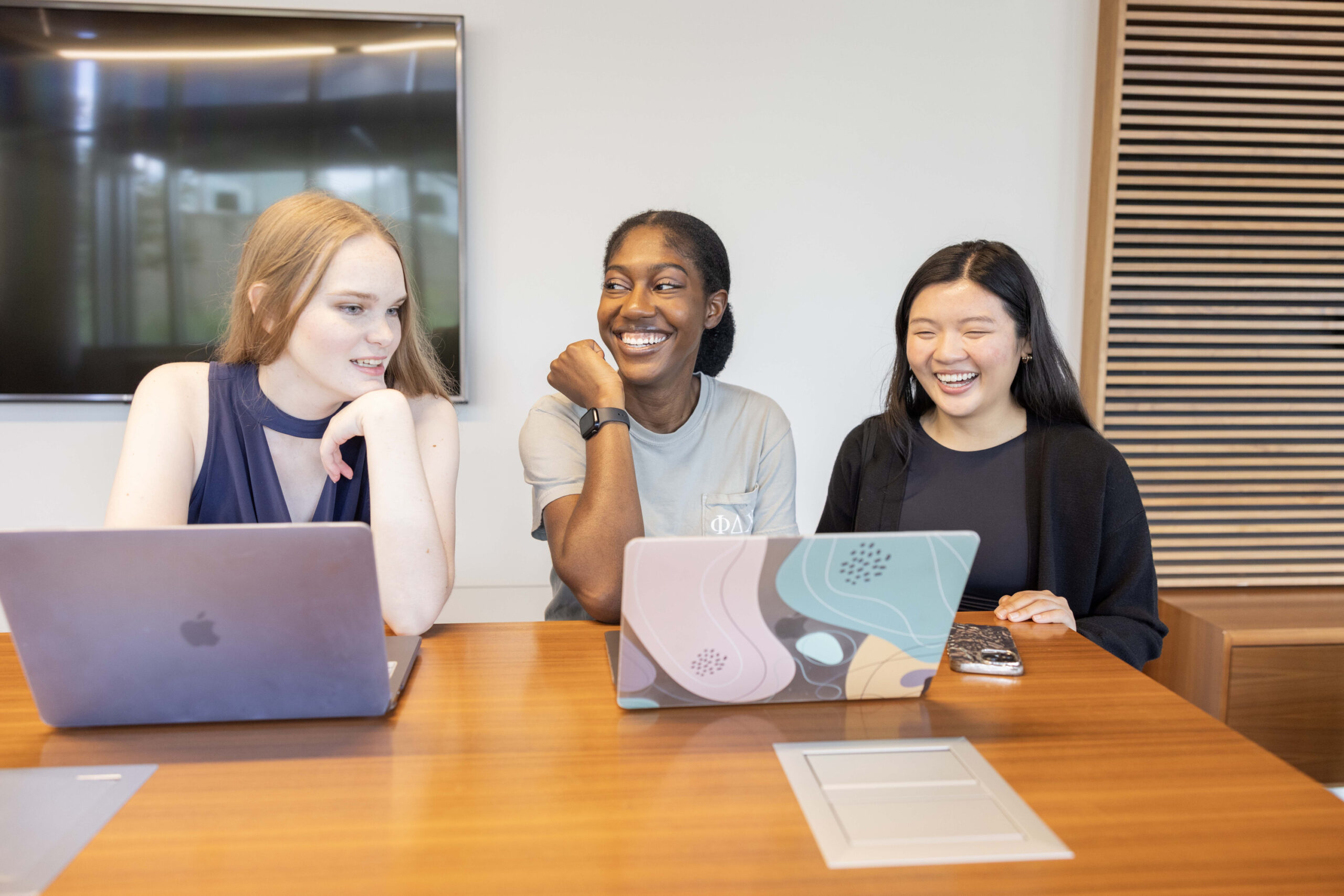 Photograph of three students smiling while sitting at a table working on laptops