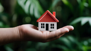 Person Holding A Miniature Red And White House In A Lush Green Setting